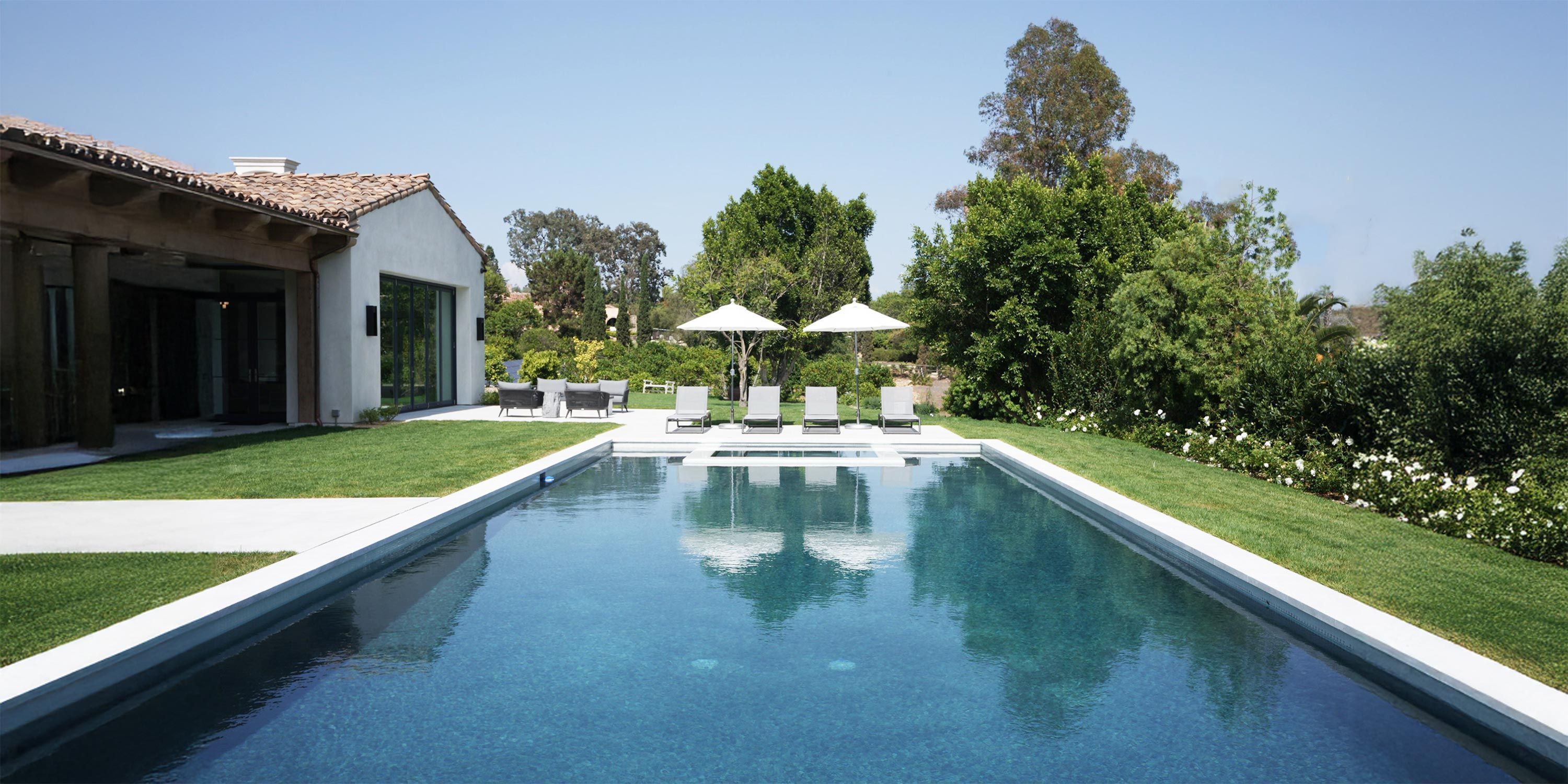 A serene backyard pool area with lounge chairs, umbrellas, and a lush green lawn framed by trees.