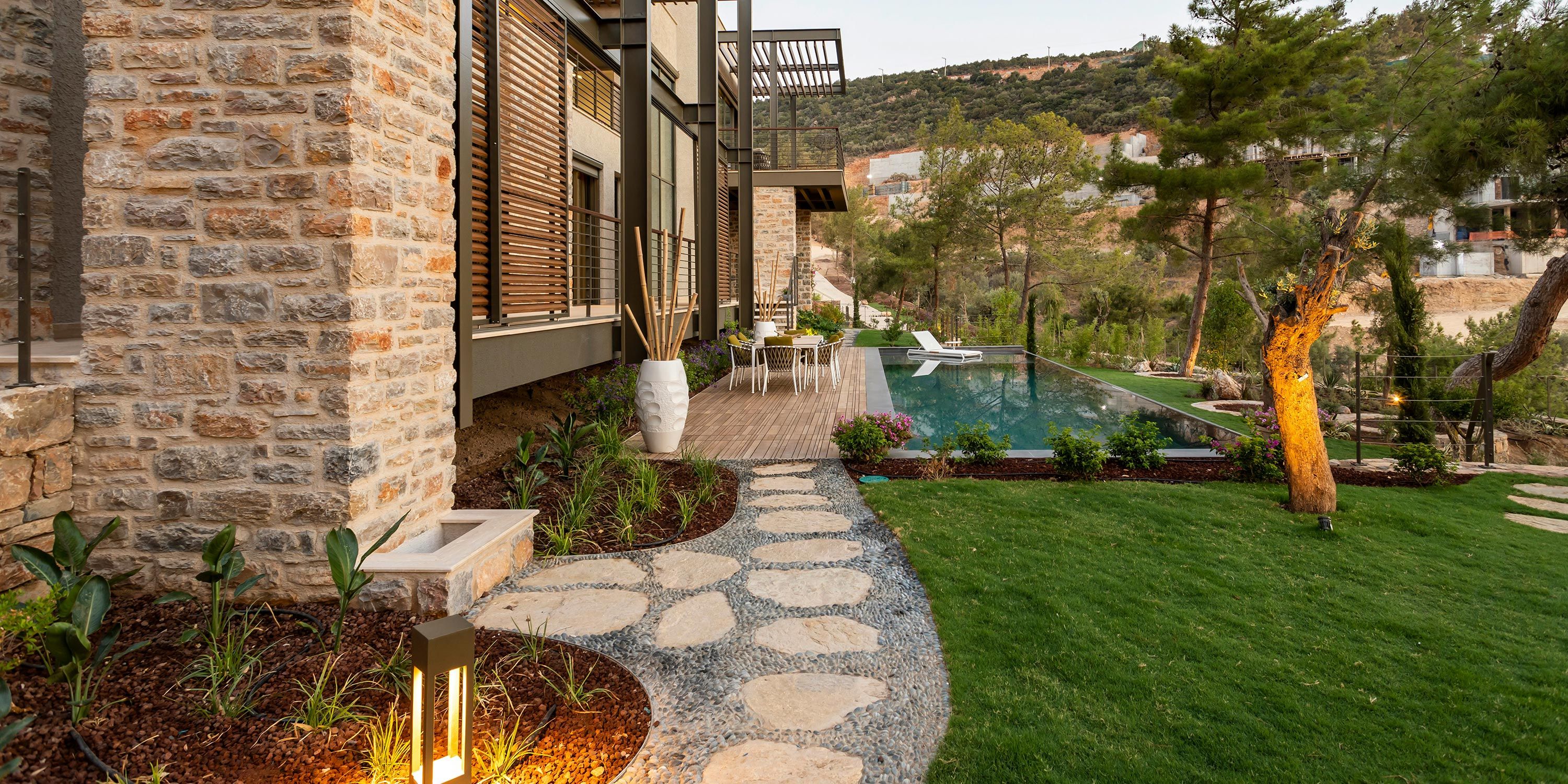 A beautifully landscaped backyard featuring a stone path, vibrant greenery, and a poolside deck with outdoor seating.