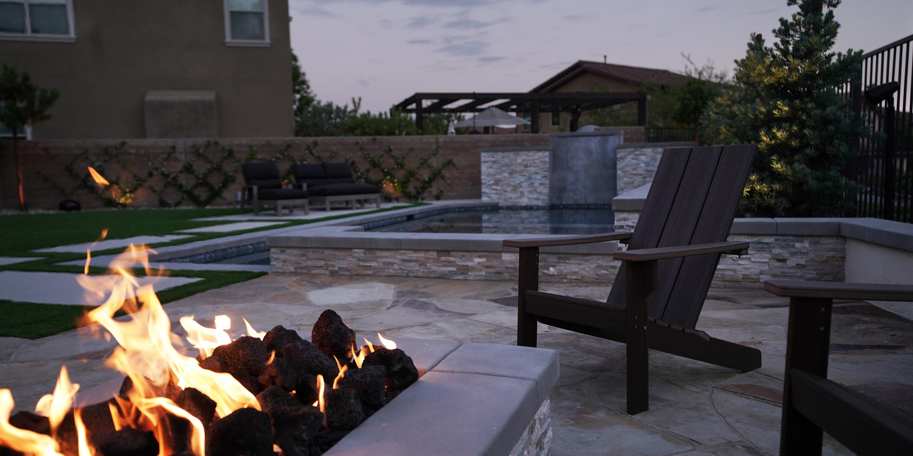 A serene outdoor seating area with a fire pit in the foreground and a pool with a stone waterfall feature in the background.