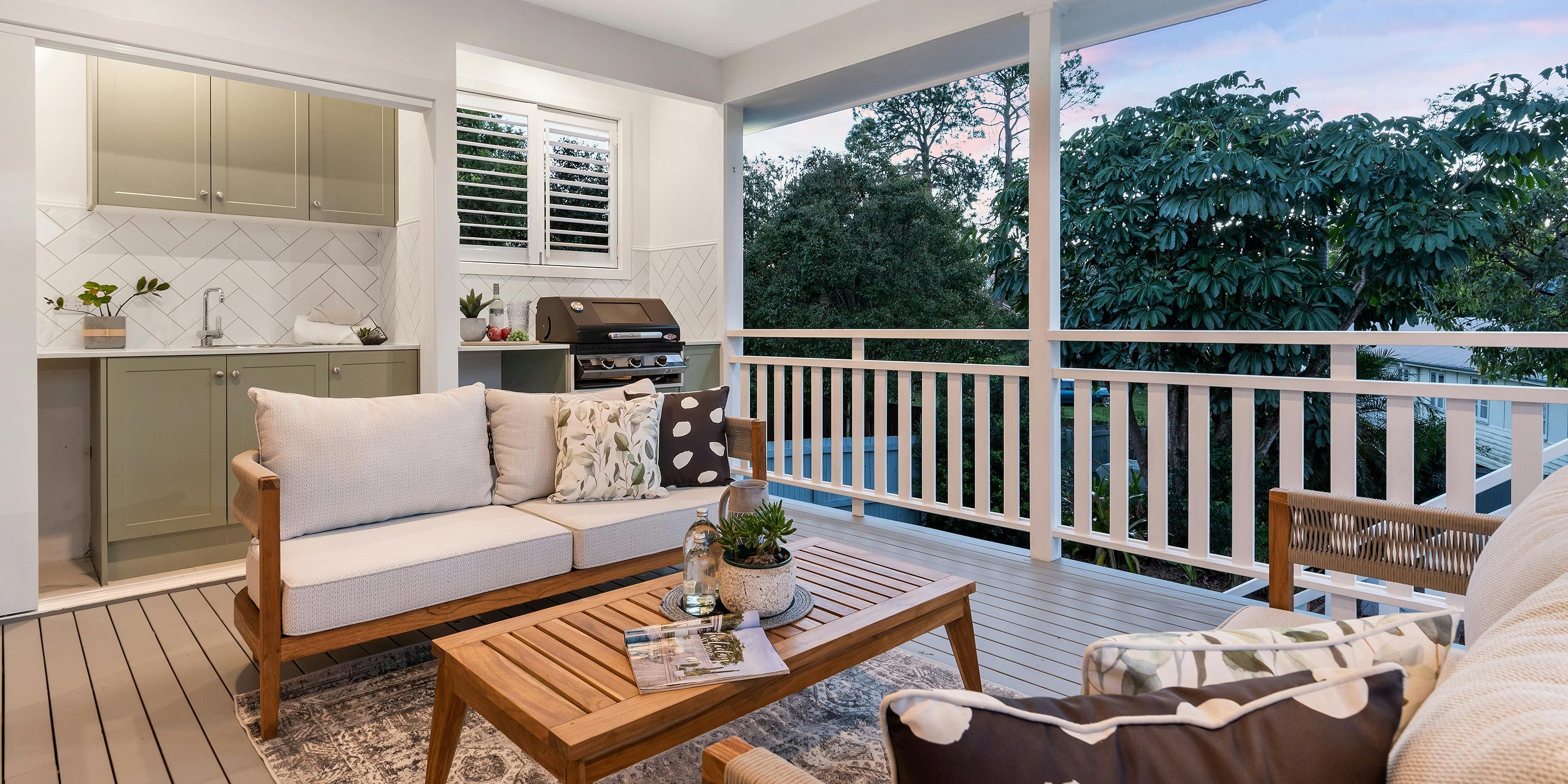 A cozy covered porch with a stylish seating area, a wooden table, and a built-in grill overlooking a lush garden.