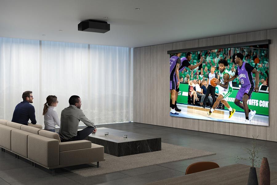 A modern minimalist home theater with a large projection screen displaying a basketball game, with three people seated on a simple beige couch.