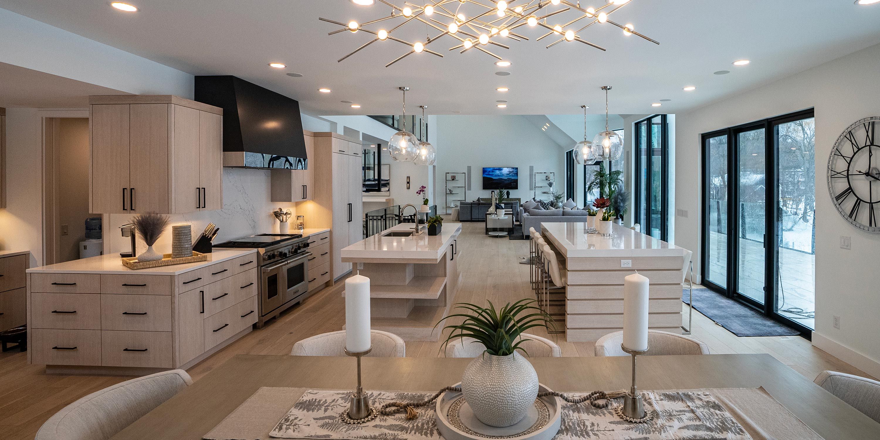 A large kitchen with a dining area featuring modern furniture and light fixtures, leading into an open-plan living space with expansive windows.