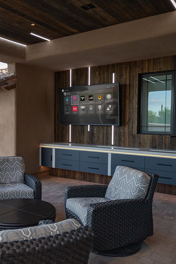 An outdoor living area with a mounted smart home interface display, comfortable seating, and rustic wood paneling.